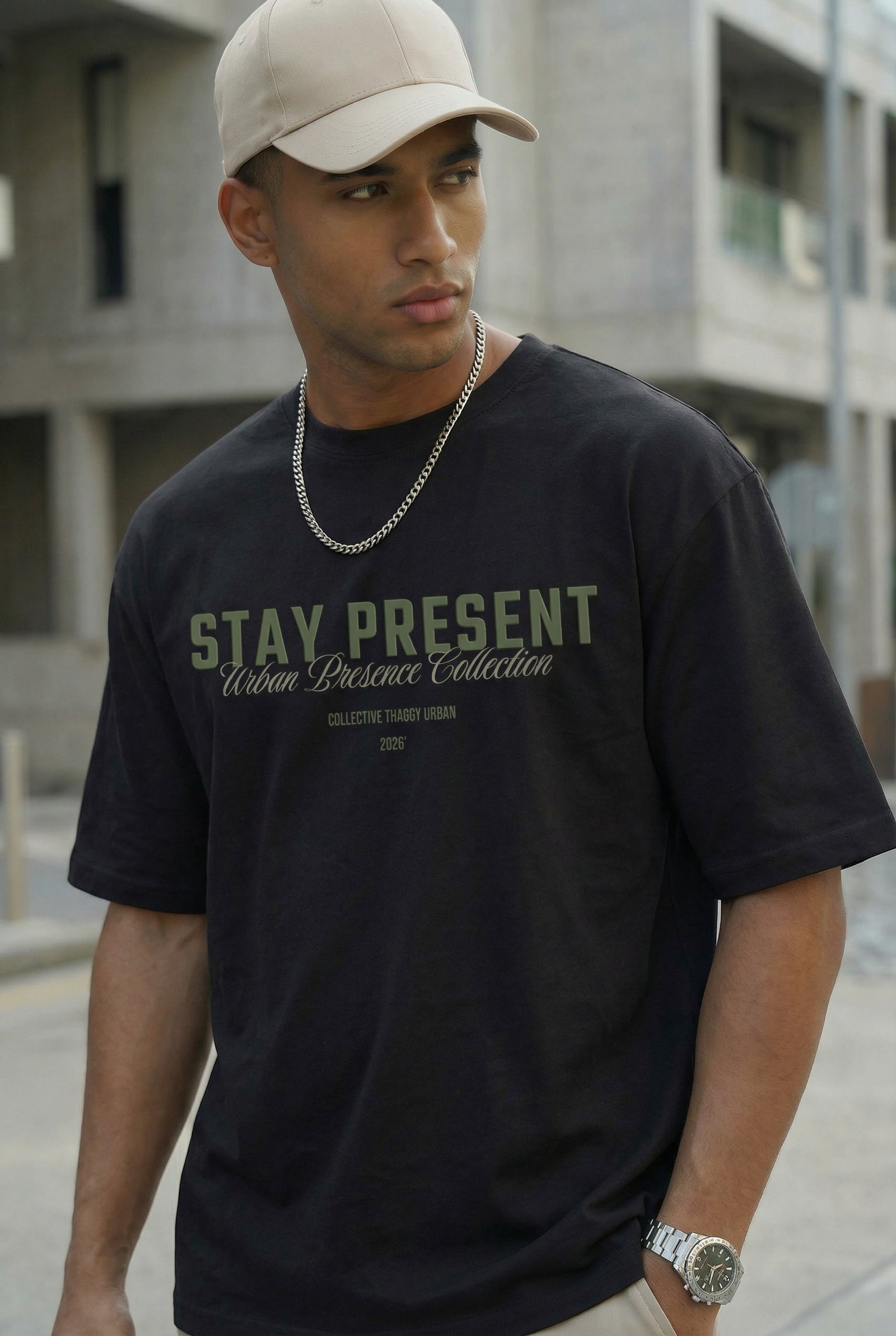imagem de um homem negro em um ambiente urbano vestindo uma camisa oversized com a estampa "stay present" da thaggy, na cor preta, ele usa corrente, boné e relógio caracteristicos o estilo streetwear.